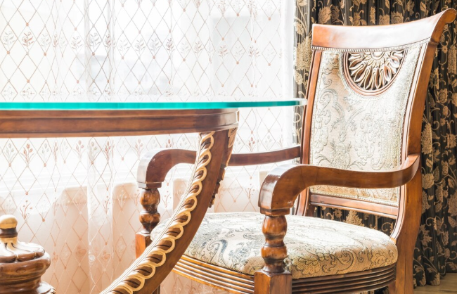 Elegant wooden chair with carved details next to a glass table in a room with patterned curtains. Sunlight filters through a window.
