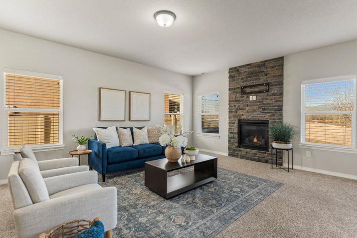 Staged open-concept living room with bold blue sofa, neutral accent chairs, fireplace, and blue patterned rug connecting to kitchen and dining areas.