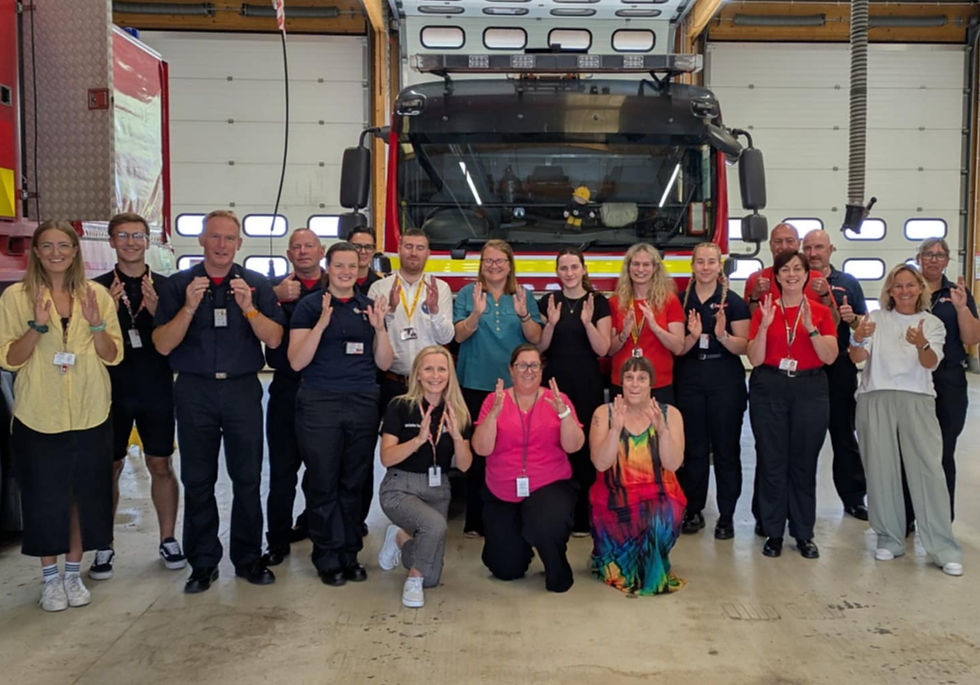 Group of people, staff members from Avon Fire & Rescue service posing in front of a fire truck inside a garage, smiling and gesturing with hands. Bright uniforms and casual attire.