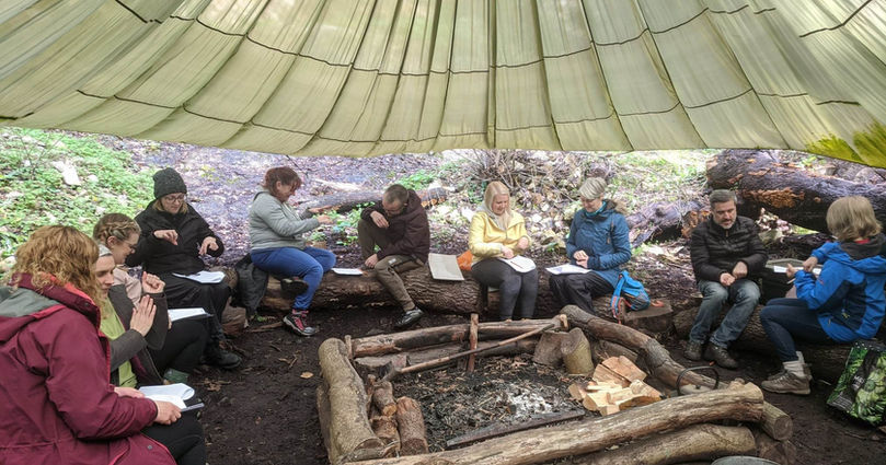 Playwild CIC outdoor practitioner setting, people are sitting round a campfire on large logs, under an old army parachute, practising the Makaton signs they have learnt during the session.