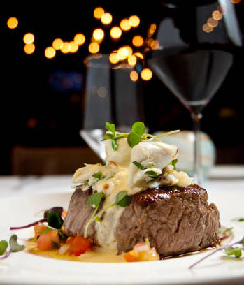 Gourmet steak dish served with vegetables and sauce on a white plate, with a glass of red wine in the background.