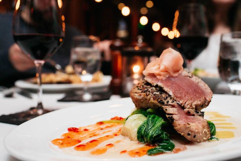 Elegant plated dish featuring seared tuna with vegetables and sauce, served at a fine dining restaurant with wine glasses in the background.