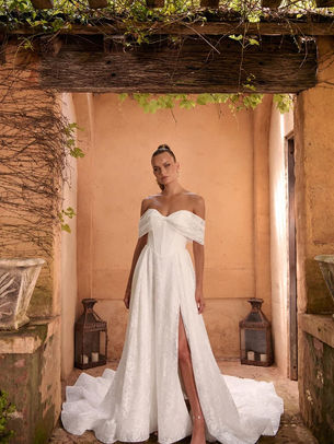 Westyn, a delicate chantilly lace gown, comprising softness and texture without compromising its clean sophistication. The off-shoulder, twist-front neckline evokes the shape of a bow. An effortlessly romantic detail that adds a hint of sweetness to the structured bodice.

Exposed corset boning offers a contemporary contrast to the gown’s otherwise timeless design. The Basque waist enhances and defines the figure, creating a beautifully cinched silhouette. The full A-line skirt flows with subtle volume, striking the perfect balance between structure and softness.

Toward the back, Westyn carries a sweeping train adorned by a row of vertical buttons trailing from the bodice to the hem.

Also available without a skirt split
