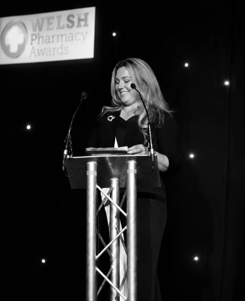 Black-and-white photo of a smiling woman speaking at a lectern beneath a “Welsh Pharmacy Awards” sign, creating a formal, celebratory mood.