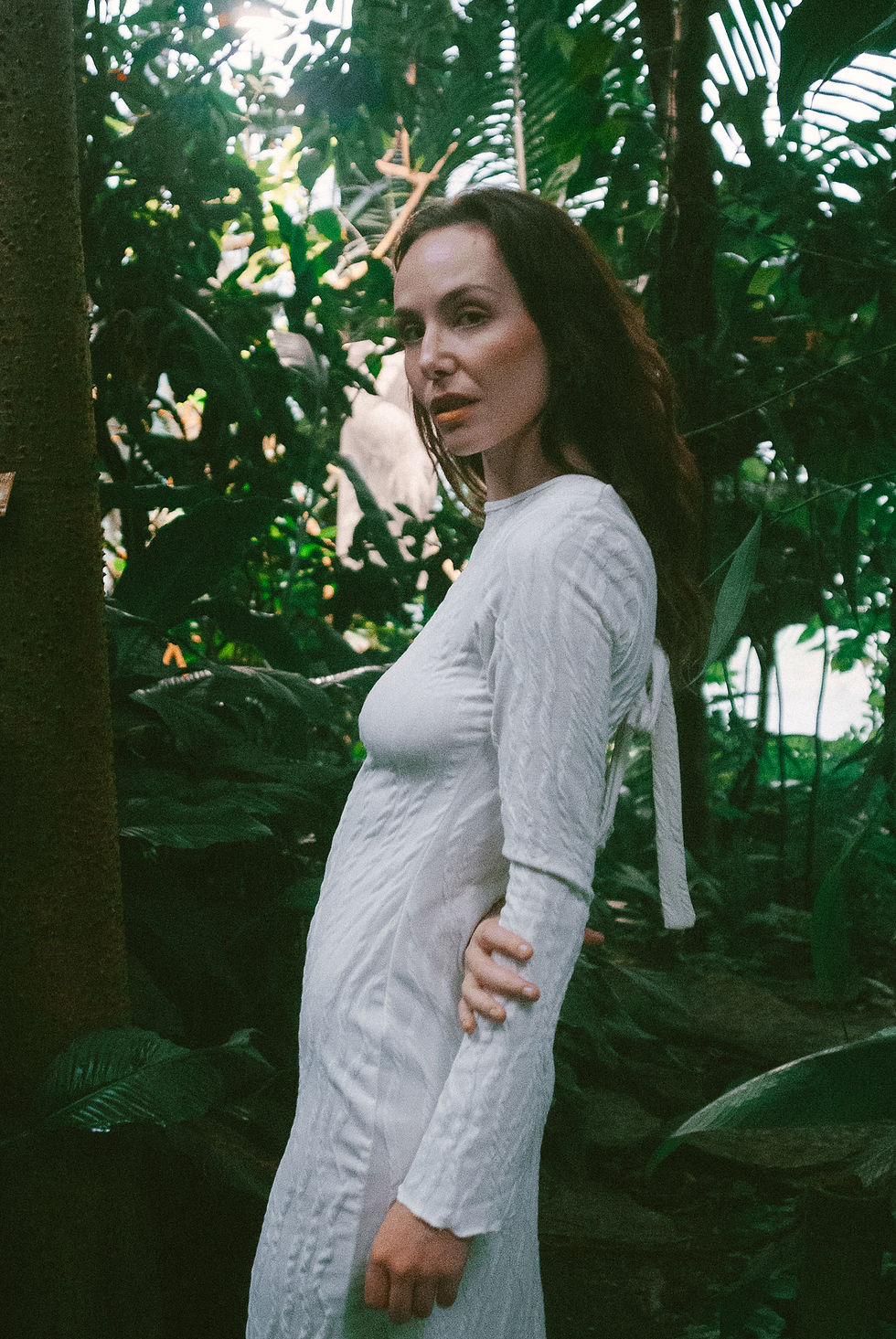 Charleen Kloss poses in profile among dense tropical vegetation while wearing a fitted white long-sleeved dress. Soft natural light filters through the greenhouse canopy, giving the image a moody, organic quality.