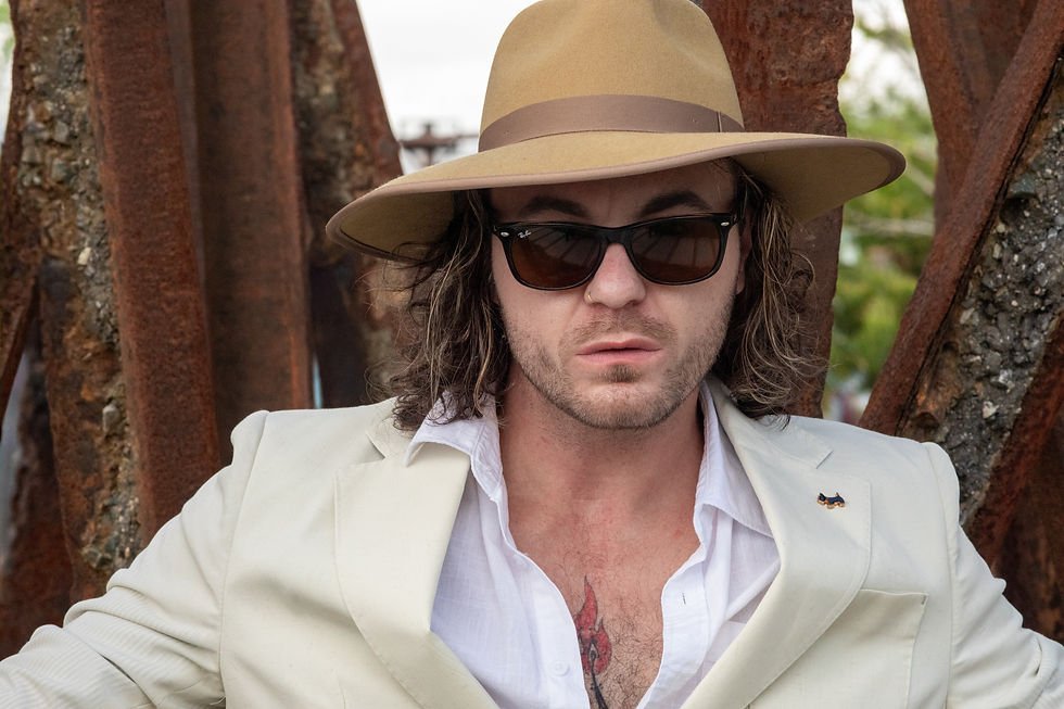 Close-up portrait of Bradley Setzer wearing a tan wide-brim hat, sunglasses, cream blazer and open white shirt while framed by rusted industrial beams. The intimate composition mixes polished style with the raw textures of Camp North End.