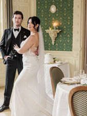An elegant wedding reception at The Inn at Erlowest features a tablescape of white and ivory florals, greenery, candlelight, gold-rimmed place settings, and neutral linens. In the softly focused background, a bride and groom share an intimate moment amid the historic venue’s distinctive green-and-gold interiors. Photographed by Rafael Gonzalez.