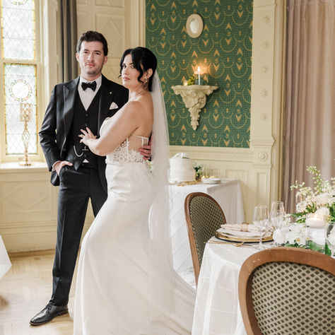An elegant wedding reception at The Inn at Erlowest features a tablescape of white and ivory florals, greenery, candlelight, gold-rimmed place settings, and neutral linens. In the softly focused background, a bride and groom share an intimate moment amid the historic venue’s distinctive green-and-gold interiors. Photographed by Rafael Gonzalez.