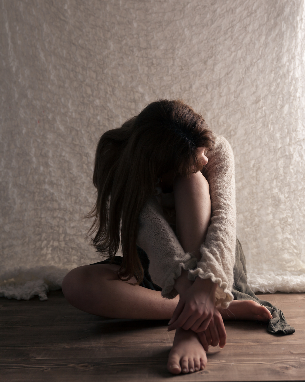 Seated woman hugs knees, head resting on arms, in a softly lit room with textured white background. Mood appears reflective or pensive.