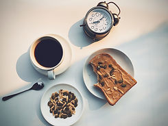 a clock, peanut butter toast on a white plate, cup of coffee, small plate of grains and nuts