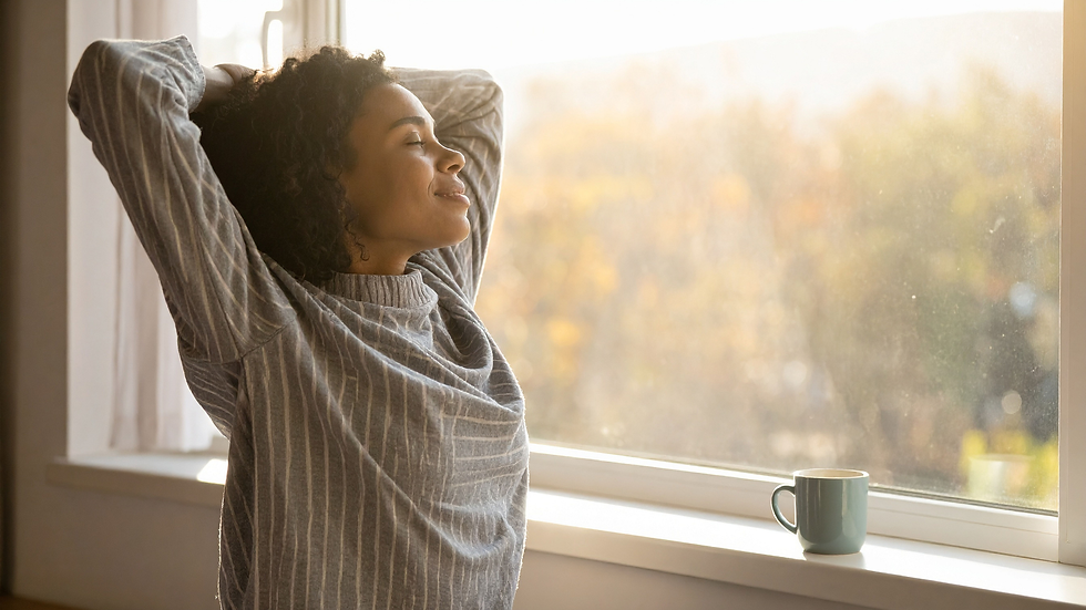 Woman in a striped sweater stretches contentedly by a sunny window. A blue mug sits on the windowsill; autumn colors blur outside.