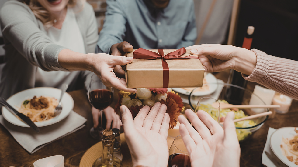 Hands exchanging a gift wrapped with a red bow over a dinner table with pasta, salad, wine and flowers. Warm, celebratory atmosphere.