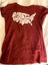 A maroon t-shirt sits on a bed. It reads "ROAD TRIP" in a font that expands the letters across in the shape of the United States