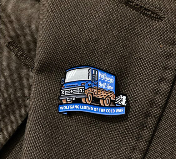 Humorous enamel pin badge featuring Wolfgang the bratwurst vendor—an iconic Cold War character beloved by BAOR veterans, complete with bratty cart and cheeky grin.