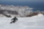 Skiier performs a deep carve on Otaru Tenguyama, with a beauitful sea view of Otaru in the background