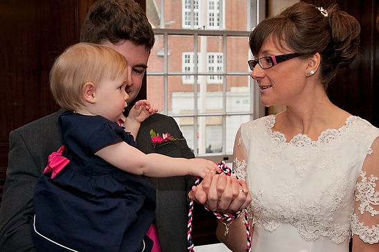 Bride in white wedding dress and groom in suit with pink buttonhole, holding their baby daughter during handfasting as part of their wedding ceremony by Tanya Jones Perfect Promises Sussex Celebrant and UK Celebrant