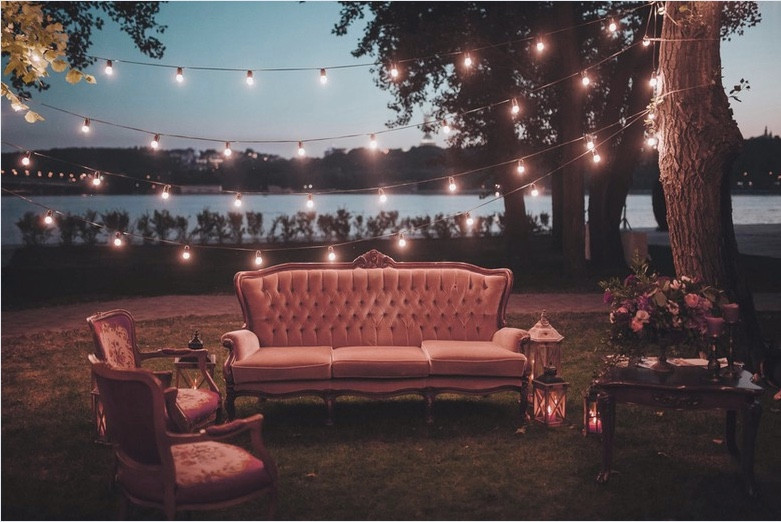 Outdoor seating area set up at outdoor wedding with plush sofa and chairs, lanterns and fairy lights strung between the trees