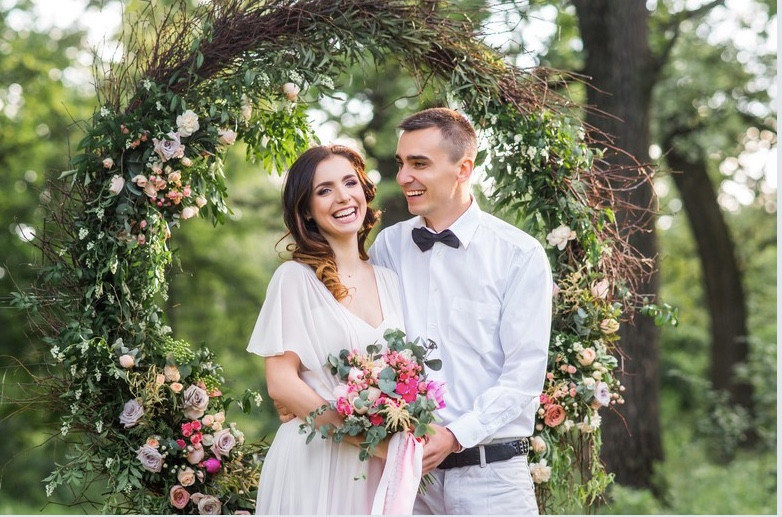 Bride in white dress with pink and white flowers in her bouquet, and groom in white shirt and black bow tie, standing in front of large hoop completely covered in flowers and foliage at their wedding ceremony.