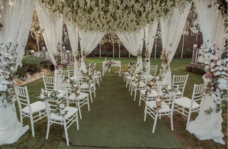 Spectacular setting for outdoor wedding ceremony for small wedding. White open-backed chairs decorated with white flowers and green foliage, white voile curtains draped around edge of ceremony area, enormous rectangular white flower cloud suspended above the length of the aisle.