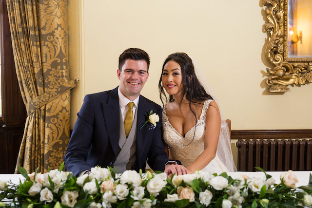 Happy bride and groom sitting at table decorated with flowers, and smiling at the camera after signing their marriage certificate
