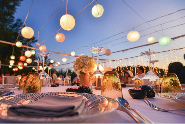 Table set for outdoor wedding reception at dusk, with amber glassware and lit paper lanterns suspended above the table