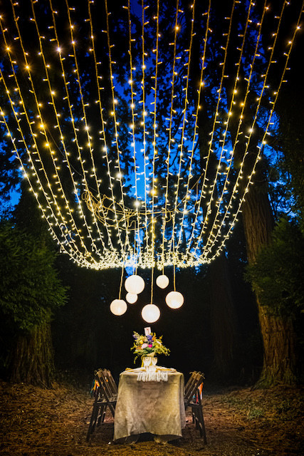 Outdoor table set for romantic dinner at dusk with canopy of fairy lights above and round lanterns hanging over the table