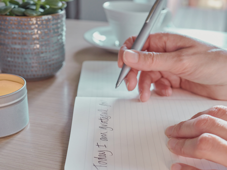 A person writing in a journal on a desk, with a candle, a plant and a cup