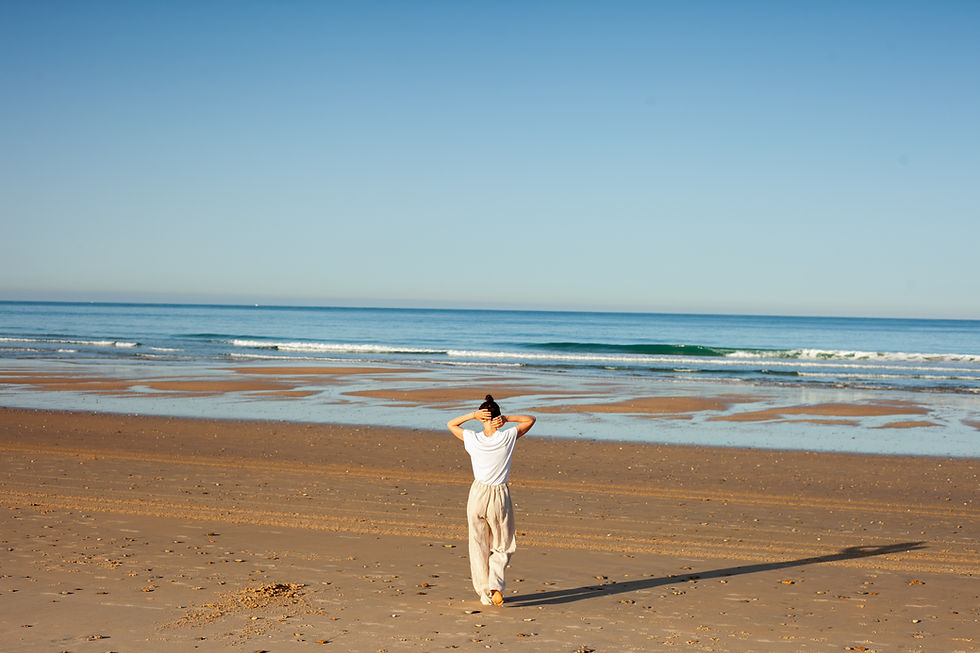 Person in Weiß steht am leeren Strand und blickt aufs ruhige Meer unter blauem Himmel.