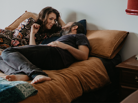 A couple relaxes on a bed with brown pillows; the woman wears a floral dress, smiling at the man in a striped shirt. Cozy room setting.