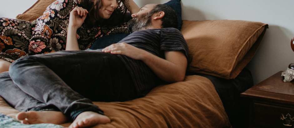 A couple relaxes on a bed with brown pillows; the woman wears a floral dress, smiling at the man in a striped shirt. Cozy room setting.