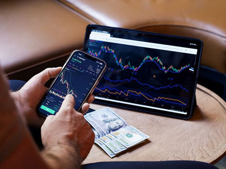 A person using a smartphone and tablet for stock market analysis, with graphs and cash visible.