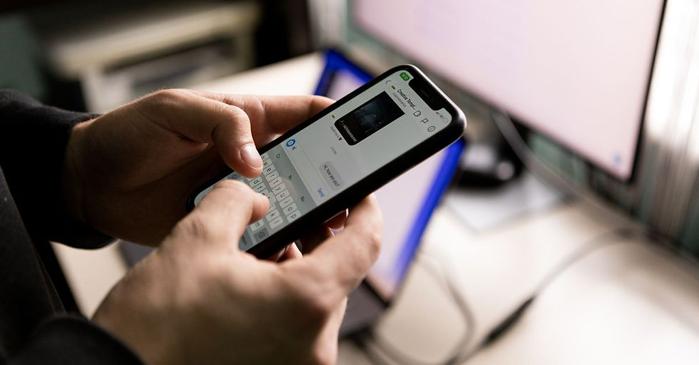 A person holding a smartphone and chatting indoors near a computer screen.