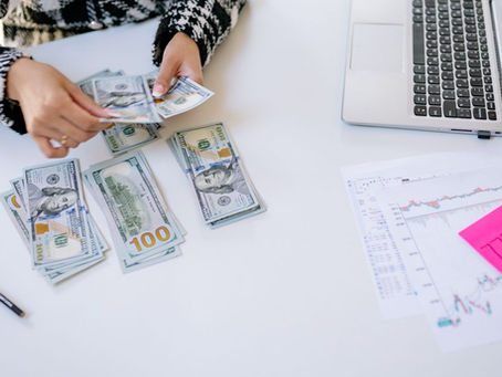 Person counting cash next to laptop and stock market charts on a white table.