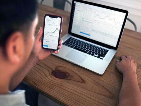 Individual reviewing stock market trends using a laptop and smartphone at a wooden table.