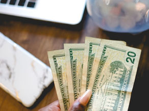 Close-up of a hand holding dollar bills beside a laptop and piggy bank, symbolizing savings and finance.