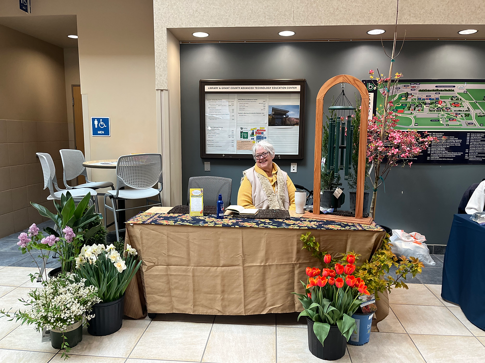 Smiling woman wearing a yellow shirt and tan vest sitting at a booth surrounded by flowers.
