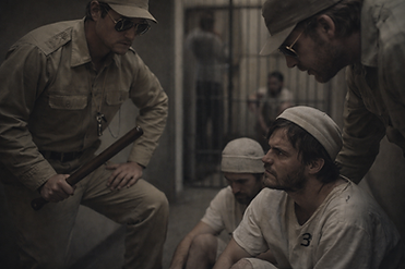 In einem düsteren, beengten Raum sitzen zwei erschöpfte Männer in einfacher Gefangenenkleidung am Boden, während zwei uniformierte Wärter mit Sonnenbrillen und Schlagstock bedrohlich über ihnen stehen; Gitterstäbe im Hintergrund und gedämpftes Licht erzeugen eine beklemmende Atmosphäre von Macht, Unterwerfung und psychischem Druck.