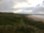Coastal landscape with grassy dunes, rocky shoreline, and sandy beach under a cloudy sky. A serene and natural scene.