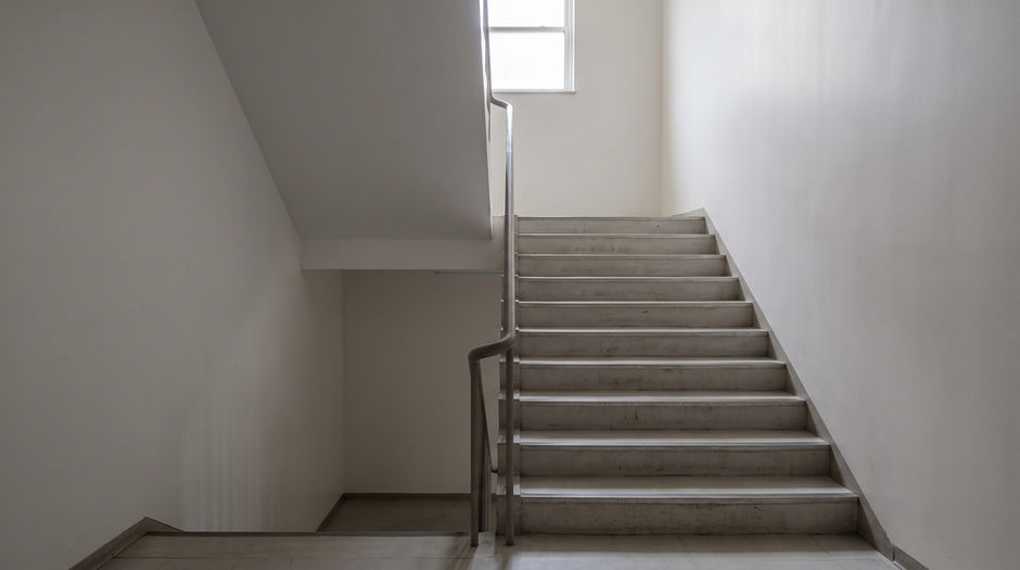 An empty institutional stairwell photographed from the bottom landing, with bare unmarked walls, a plain slim metal handrail and daylight from a single window on the upper landing.