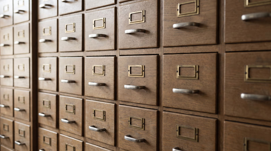 A tall bank of oak library card catalogue drawers fills the frame in raking daylight, every small brass label holder completely empty.