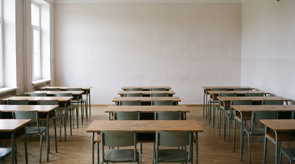 An empty secondary-school classroom seen from the back of the room, with rows of plain bare wooden desks and chairs facing a blank front wall and daylight from side windows.