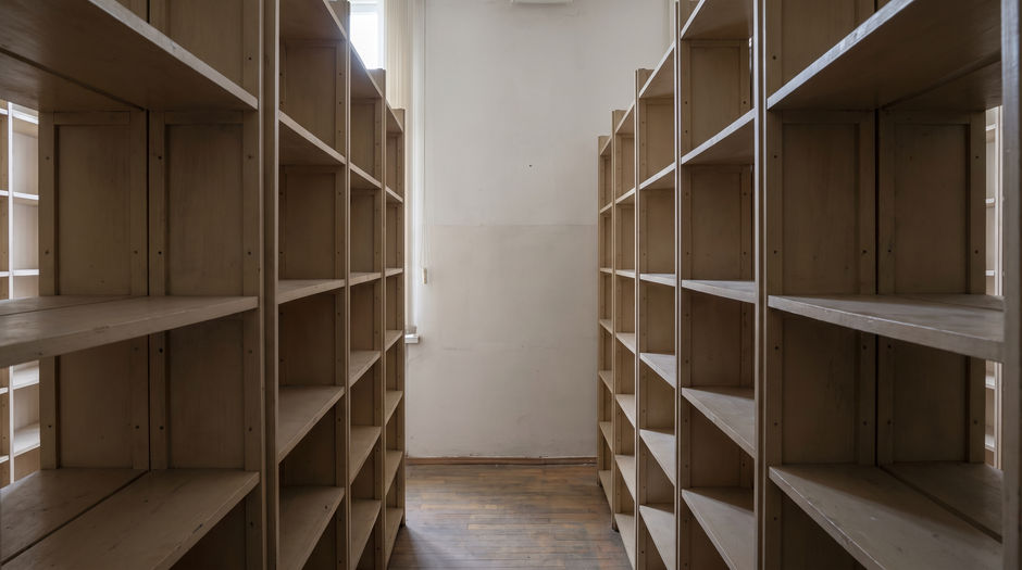 An empty library aisle seen from one end, flanked by two tall plain wooden shelving units whose shelves are completely bare and unlabelled, with a plain pale wall and a single window at the far end in soft daylight.