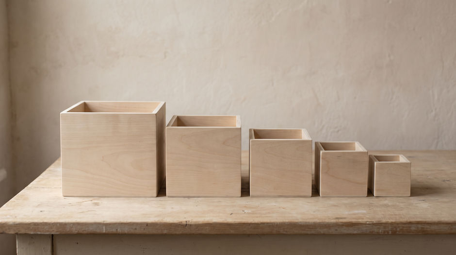 Five plain unmarked wooden boxes of graduated size arranged in a row from largest to smallest on a bare pale wooden table against a blank plaster wall.