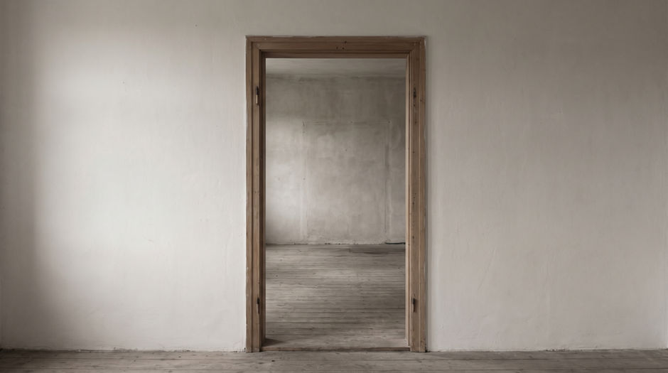 A single plain open doorway in a bare pale plaster wall, looking through into a second empty room with bare floorboards and daylight from an unseen window.