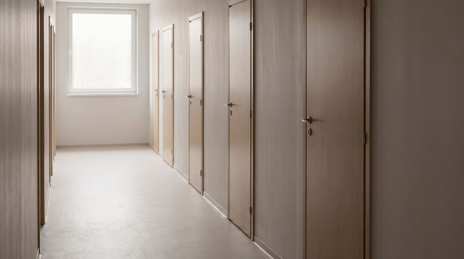 An empty institutional corridor lined with five plain closed pale wooden doors, every one of them completely blank with no nameplates, signs or numbers, lit by a single window at the far end, with no person in the frame.