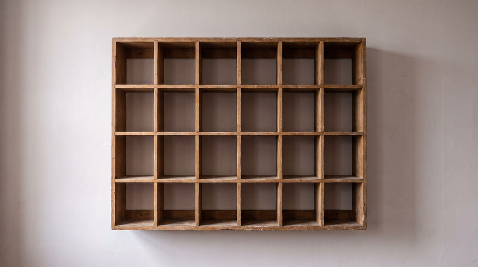 A wall-mounted wooden pigeonhole rack of twenty-four open compartments, every one of them completely empty and unlabelled, against a plain plaster wall in soft daylight.