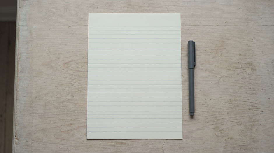 A single sheet of blank ruled writing paper lying square on a pale wooden tabletop with one plain unmarked pen resting beside it, photographed from directly above in soft daylight.