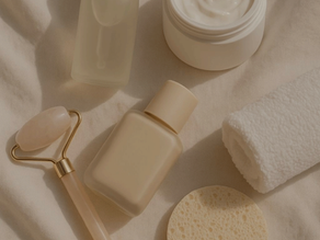 Beige background with a few bottles of skin care, lotion container open, and face roller all with no logos and higher of beige.