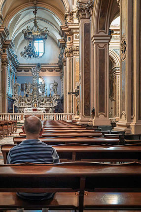 A man seated alone in the pews of an ornate church in Maglie, looking towards the high altar