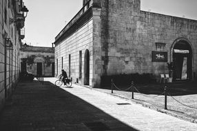 Black and white photograph of a cyclist passing a long stone building in strong sunlight in Maglie